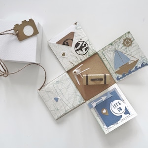Può includere: Una scatola di carta artigianale blu e marrone a tema viaggio. La scatola ha un design a mappa e presenta una bussola, un aereo, una valigia e una freccia con il testo "Let's Go".