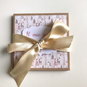 May include: A square gift box tied with a gold satin ribbon. The box features a patterned top with reindeer and Christmas trees. A white tag with the text "Il mio pr..." is visible.
