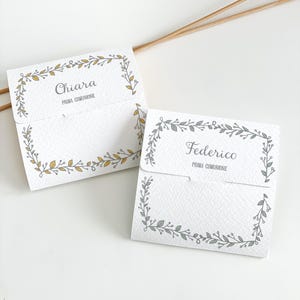 Könnte beinhalten: Zwei weiße Papierkarten mit Blattranddesigns. Eine Karte trägt die Aufschrift "Chiara" und "Prima Comunione" in grauer Schrift. Die andere Karte trägt die Aufschrift "Federico" und "Prima Comunione". Die Karten haben eine kleine Klappe und sind mit einem zarten Blattrand verziert.