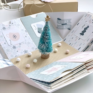 Peut inclure: Calendrier de l'Avent fait à la main, ouvert pour révéler des pochettes numérotées avec des illustrations hivernales. Un petit sapin de Noël bleu-vert avec une étoile dorée est au centre. Le texte "Winter Wonderland" est sur une bannière.