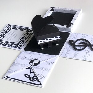May include: A black and white pop-up card featuring a miniature grand piano. The card is decorated with musical notes and a treble clef. The card also has a quote in Italian.