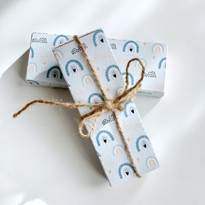 Peut inclure: Deux petites boîtes enveloppées de papier blanc avec un motif arc-en-ciel bleu, jaune et noir. Les boîtes sont liées ensemble avec de la ficelle.