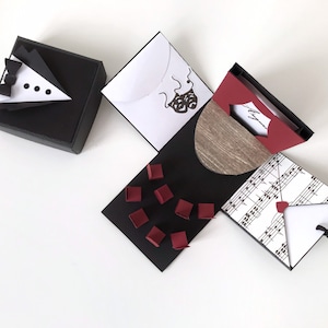 Può includere: Una scatola esplosiva in bianco e nero con un design a smoking sulla parte superiore, un design a nota musicale su un lato e un design a venatura del legno sull'altro lato. La scatola ha un interno rosso e presenta una piccola busta e diversi quadrati rossi.