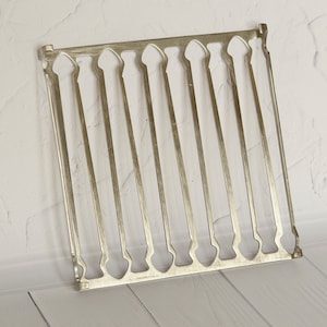 Puede incluir: Rejilla de metal plateado con un diseño decorativo. La rejilla tiene una forma rectangular y está hecha de finas barras de metal.
