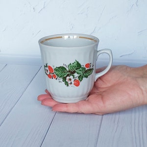 May include: White ceramic mug with a gold rim and a floral design featuring red strawberries and green leaves.