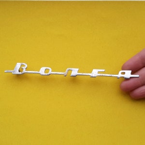 Può includere: Un emblema in metallo argentato con la parola "BONNET" scritta in lettere in rilievo.