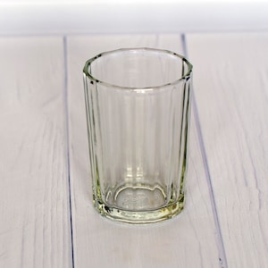 Puede incluir: Un vaso transparente con estrías y un borde ligeramente ensanchado. El vaso tiene forma cilíndrica y una base gruesa. El vaso está vacío y sobre una superficie de madera clara.