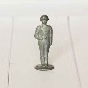 May include: A small, silver-colored metal figurine of a soldier in uniform. The figurine has a detailed design, including a helmet, uniform, and boots. The soldier is standing on a small, round base. The background is a white wooden surface.