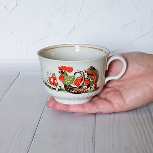 May include: A white ceramic teacup with a handle, featuring a colorful rooster illustration. The rooster is depicted with red, green, and brown details, along with strawberries. The cup has a gold rim and is held in a hand.