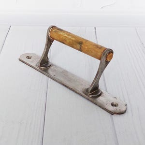May include: A vintage door pull featuring a wooden handle and a metal base. The handle is light brown, and the metal base has a silver finish. The pull has a rectangular shape with rounded edges, designed for easy installation, and is suitable for various doors.