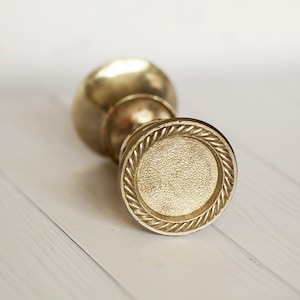 Peut inclure: Bouton de porte en métal doré avec un motif de corde autour du bord. Le bouton a une surface lisse et légèrement texturée.