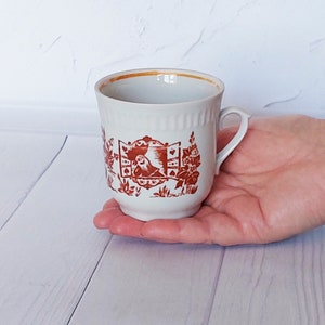 May include: A white ceramic coffee mug with a brown floral design and a gold rim. The mug has a handle and is decorated with a scene of a bird in a window.