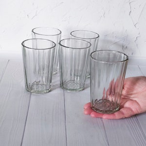 May include: A set of five clear, ribbed drinking glasses. One glass is held in a hand, while the others are arranged on a light-colored wooden surface. The glasses have a simple, classic design.