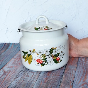 Puede incluir: Olla de esmalte blanco con tapa, con un diseño de flor de cerezo con cerezas rojas, flores blancas y hojas verdes. La olla tiene un asa en la tapa y dos asas laterales. Está sobre una superficie de madera.