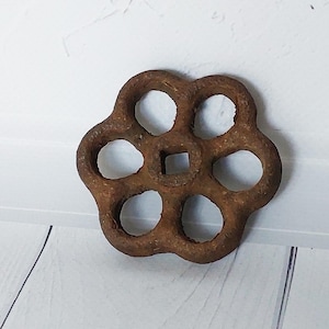 Könnte beinhalten: Ein rostbrauner, blütenförmiger Ventilkurbel mit sechs kreisförmigen Öffnungen. Die Mitte hat eine quadratische Öffnung. Das Metall weist Gebrauchsspuren auf und hat eine strukturierte Oberfläche. Wahrscheinlich ein Wasser- oder Gasventilgriff.