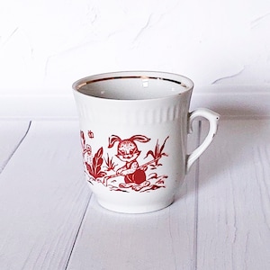 May include: White ceramic mug with a red and gold rim. The mug features a red illustration of a rabbit holding a rake in a garden setting.