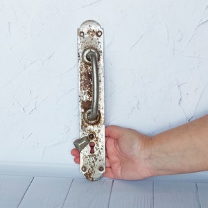 May include: An antique door handle with a distressed, white finish and visible rust. The handle has a curved, silver-colored grip and a keyhole. The overall design is rectangular with rounded edges and mounting holes.