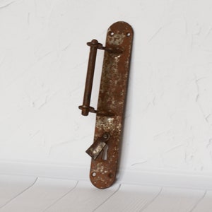 Op de afbeelding: Een roestige metalen deurknop met een lange, rechthoekige plaat en een hendel. De knop heeft een sleutelgat en een klein, decoratief label.