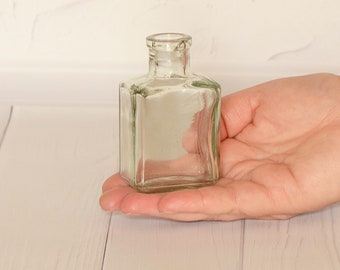 May include: A clear glass bottle with a square shape and a rounded top. The bottle is empty and has a slight green tint.