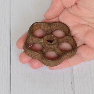 May include: A rusty, brown metal valve handle with a flower-like design. The handle has five circular openings and a central hexagonal socket. The aged appearance suggests it is vintage or antique.