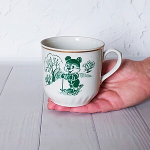 以下が含まれることがあります： 緑色の冬景色の中でスキーをしているクマのイラストが描かれた白いセラミック製のマグカップ。マグカップは金色の縁取りが施されています。