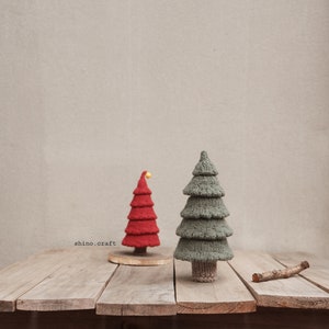 Könnte beinhalten: Zwei gestrickte Weihnachtsbäume auf einer Holzoberfläche. Der größere Baum ist grün und der kleinere Baum ist rot mit einem gelben Stern oben. Der Text "shino.craft" ist in der linken unteren Ecke des Bildes sichtbar.