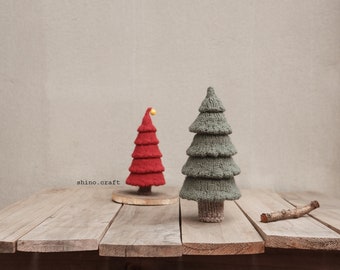 May include: Two knitted Christmas trees on a wooden surface. The larger tree is green and the smaller tree is red with a yellow star on top. The text "shino.craft" is visible in the lower left corner of the image.