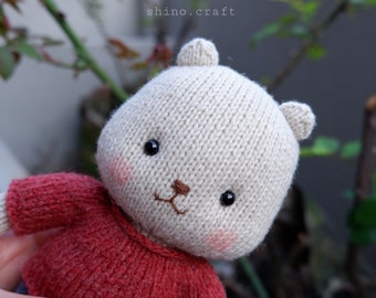 May include: A knitted plush toy bear wearing a red sweater and blue jeans. The bear has a beige body, black eyes, and a brown nose.