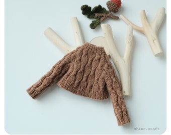 May include: A brown knitted sweater with a cable knit pattern hangs on a wooden branch. The sweater is a small size, suitable for a doll or small child.