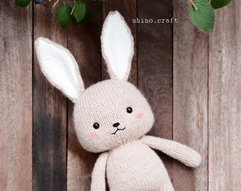 May include: A beige knitted bunny rabbit toy with white ears and black eyes. The bunny is wearing green knitted booties and is lying on a wooden surface.