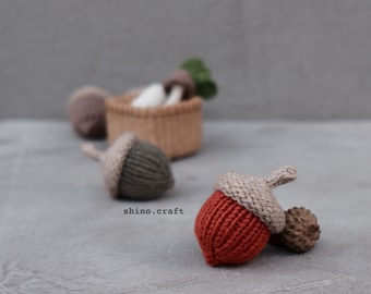 May include: Three knitted acorn ornaments, one red, one brown, and one gray, with a brown stem on each. The acorns are sitting on a gray surface. The text "shino.craft" is visible in the image.