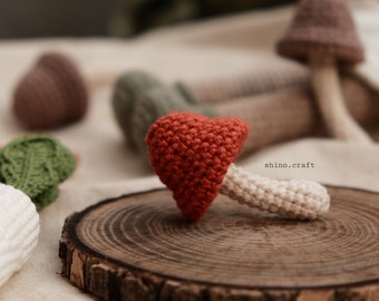 May include: A crocheted mushroom with a red cap and a white stem. The mushroom is sitting on a wooden slice. The text "shino.craft" is visible in the image.