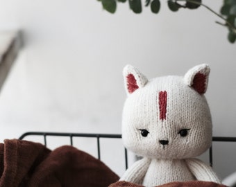 May include: White knitted plush toy cat with red accents sitting in a brown blanket. The cat has black eyes and a black nose.