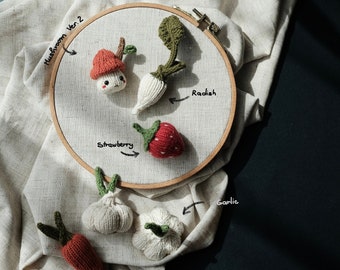 May include: A collection of knitted food items, including a mushroom, radish, strawberry, garlic, carrot, and another mushroom. The items are arranged on a white fabric background with handwritten labels identifying each item.
