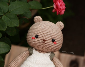 May include: A crocheted brown bear wearing a white dress with green straps. The bear has black eyes and red cheeks. The bear is sitting on a wooden surface with a pink rose in the background.