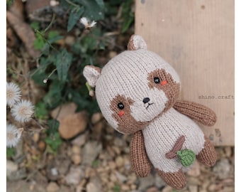 May include: A knitted raccoon plush toy with brown and beige fur, black eyes, and a small green leaf on its belly. The toy is sitting on a bed of brown rocks and green plants.