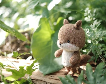 May include: A brown and white knitted squirrel toy with a long tail, sitting on a wooden surface with green foliage in the background.