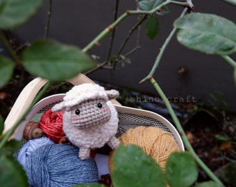 May include: A white crocheted sheep amigurumi toy with black eyes and a red nose sits in a basket with balls of yarn in blue, red, and yellow.
