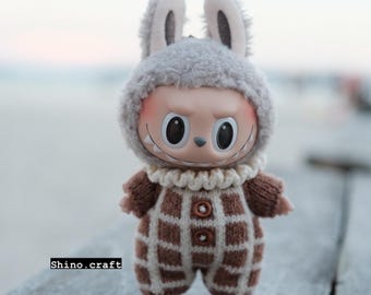 May include: A doll wearing a gray bunny hat and a brown and white checkered knit outfit. The doll has a smiling face with large eyes and is standing on a wooden surface. The text "Shino.craft" is visible.