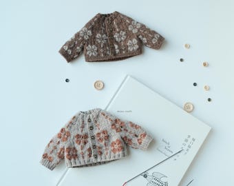 May include: Two brown and beige knitted cardigans with floral patterns and brown buttons. The cardigans are laying on a white surface next to a book with Japanese text and a red knitting needle.