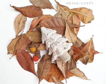 May include: A white knitted Christmas tree with colorful accents sits on a bed of brown autumn leaves. Two small crocheted acorns are also visible in the image.