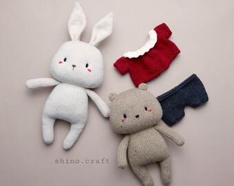May include: Three knitted toys: a white bunny, a brown bear, and a dark blue pair of pants. The bunny has red cheeks and a white collar. The bear has red cheeks and a brown nose. The pants have a white trim.