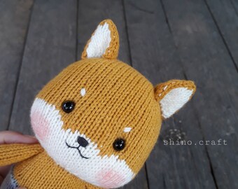 May include: A knitted plush toy dog with brown fur, white ears, and black eyes. The toy has a pink nose and a white mouth.
