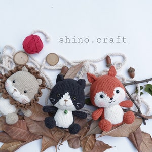 May include: Three knitted stuffed animals: a lion, a cat, and a fox. The lion is light brown with a brown mane, the cat is black and white, and the fox is orange with white accents. The animals are surrounded by fall leaves, a small tree, and twine.