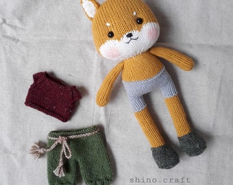 May include: A knitted fox doll with a gray and white outfit, a maroon knitted shirt, and green knitted pants with a brown belt. The doll is lying on a beige fabric background. The text "shino.craft" is visible in the bottom right corner of the image.