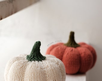 May include: Two knitted pumpkins, one white with a green stem and one orange with a green stem. The pumpkins are sitting on a white surface. The text "shino.craft" is visible in the background.