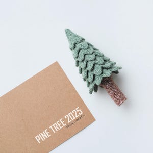 Peut inclure: Une décoration de sapin de Noël en tricot vert avec un tronc brun. L'ornement est posé sur une carte en papier brun avec le texte "PINE TREE 2025 shino.craft".
