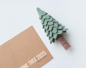 May include: A green knitted pine tree ornament with a brown trunk. The ornament is sitting on a brown paper card with the text "PINE TREE 2025 shino.craft".