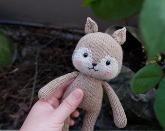 May include: A handmade knitted fox doll in shades of tan and white. The fox has a sweet expression with rosy cheeks, black eyes, and a small black mouth. The doll has small ears and is holding its arms out. The doll is approximately 8 inches tall.
