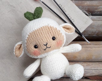 May include: A white knitted lamb with a green leaf on its head. The lamb has black eyes and pink cheeks. The lamb is sitting on a wooden surface with knitting needles in the background.